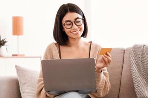Shopping On Internet From Home, Ecommerce Concept. Smiling young asian woman sitting on couch at home, ordering and buying clothes or products online, paying with credit card and using laptop photo