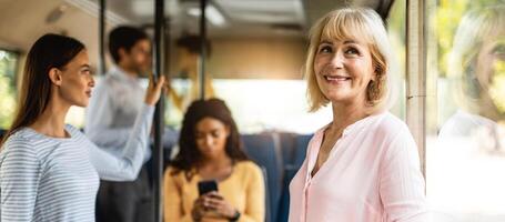 Public Transportation Concept. Portrait of smiling senior lady standing in bus, leaning looking out of window, going home, banner, panorama. Group of diverse people passengers in blurred background photo