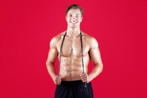Handsome young sportsman with bare torso and jumping rope looking at camera and smiling on red studio background. Shirtless millennial guy ready for cardio workout or aerobic exercises photo