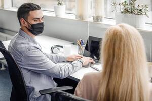 Modern workplace in coworking office after covid quarantine. Guy and lady in protective masks sitting at table and working on laptop, talking, through protective glass in interior, panorama, empty space photo