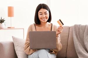 Online Shopping And Delivery Concept. Happy asian woman making purchases on internet with laptop, holding and showing credit card to pay. Smiling lady using computer sitting on sofa, copy space photo