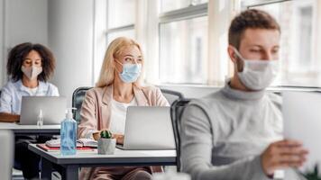 Teamwork on project. Multiracial employees in protective masks work on laptops at desks with antiseptics, focus on pensive blonde lady, looking out the window, coming up with idea, copy space photo