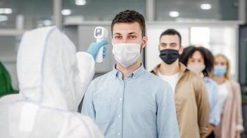 Modern technologies for determining temperature with non-contact thermometer. Man in hazmat suit measures temperature of multiracial colleagues in protective masks at entrance to office, free space, indoors photo