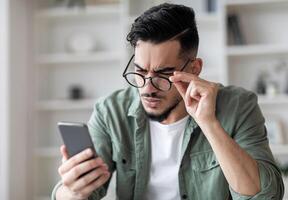 Eyesight Problems Concept. Young Arab Man In Eyeglasses Looking At Smartphone Screen And Frowning, Millennial Guy Trying To Read Message, Suffering From Astigmatism And Bad Vision, Closeup Shot photo