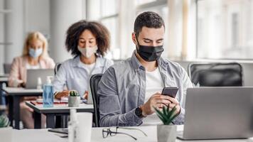 Modern gadgets for working with clients remotely from office after quarantine. Arabic young guy in protective mask typing on smartphone at table with laptop in office interior photo