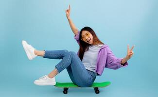 Joyful asian lady sitting on skateboard with legs up and smiling to camera, showing v-sign gesture. Young woman spending time and having fun over blue studio background photo