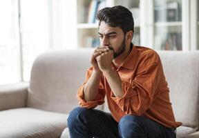 Permacrisis, life problems, financial hangout concept. Pensive young middle eastern man sitting on sofa at home, leaning on his hands, experiencing difficulties in life, looking at copy space photo