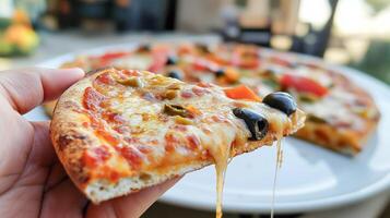 A hand holds a slice of cheesy pizza topped, with vegetables and olives as the rest of the pizza sits on a plate in a warm outdoor environment enjoying a meal. photo