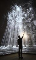 Silhouette of a man reaching towards projected digital architecture in a dark room photo
