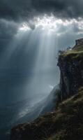 Dramatic sunbeams illuminate a rugged cliffside landscape with moody clouds and distant mountains photo
