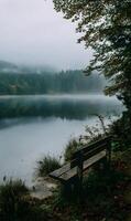 A tranquil scene unfolds with a wooden bench overlooking a misty lake and a serene forest photo