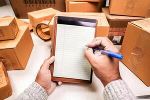 Seller Checking Packing boxes, Hands with pen writing on a blank notepad near cardboard boxes. Inventory check or packing list creation. e-commerce concept photo