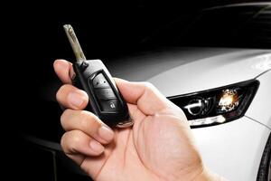 Car Key Fob, A hand holds a car key with a blurred white car in the background. vehicle access, car remote. photo