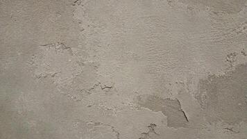 Close-up of a rough, worn-out concrete wall with a gritty texture, featuring cracks and crevices. The color is a dull, muted gray with subtle variations in shade, showing off the uneven surface. photo