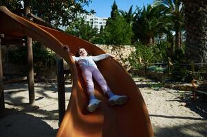 Smiling Child Sliding on Playground Slide Amid Green Palm Trees and Stone Walls photo