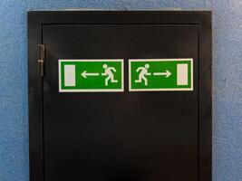 Two emergency exit signs with running figure and arrows pointing left and right placed on black door panel against blue wall, providing clear direction for safe evacuation in case of emergency photo