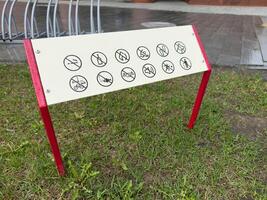 Prohibition sign with multiple black icons on a white board supported by red legs, installed on a lawn next to a walkway, indicating banned activities in a public space for safety and order photo