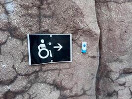 Accessibility sign with wheelchair icon and arrow mounted on textured stone wall next to service button, indicating direction and call support for people with limited mobility in public environment photo