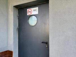 Black door with round frosted window and no smoking WC sign mounted on rough gray wall, marking entrance to public restroom with emphasis on clarity, hygiene and behavioral regulation in urban setting photo