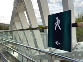 Close up of pedestrian walkway sign with walking figure and arrow on modern glass and steel bridge, with urban buildings and green trees visible in the background photo