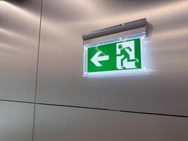 Emergency exit sign with left arrow and running man symbol mounted on metal wall. Green illuminated panel used for evacuation guidance in public building. Concept of safety, escape and compliance photo
