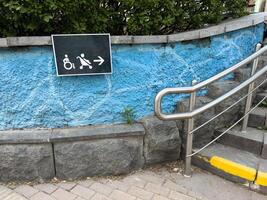 Accessibility sign with icons of wheelchair and baby stroller directing right, mounted on textured wall next to ramp with metal railing, promoting inclusive design in urban pedestrian environment photo