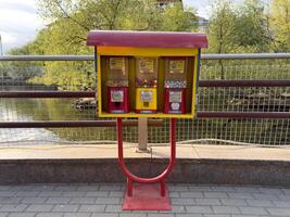 Colorful candy and gum vending machine stands on paved riverside path, inviting passersby to enjoy sweet treat. Surrounded by greenery, adds playful touch to the urban scene photo