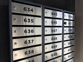 Row of modern stainless steel mailboxes with bold black numbers and built in locks mounted on dark wall in apartment building lobby, illustrating secure personal storage in urban residential space photo