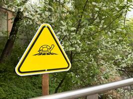 Bright yellow triangular warning sign with turtle icon stands in green park zone, serving as clear visual symbol in public space, combining environmental education with modern graphic design photo
