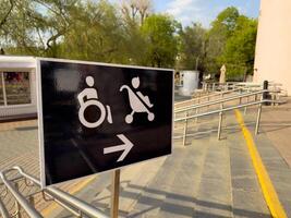 Accessibility sign showing direction to ramp for wheelchair users and strollers, located outside public building with handrails and trees in the background on sunny day photo