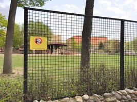 Wire fence with yellow warning sign in zoo area instructing visitors not to feed animals, surrounded by green grass, trees and enclosures, serving as protective barrier and rule enforcement element photo