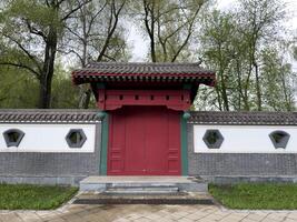 Traditional red wooden gate with tiled roof and geometric windows in decorative wall, inspired by East Asian architecture. Symmetry design and natural park background make. Ideal for visual projects photo