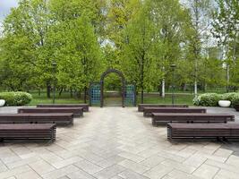 Well maintained public park area with symmetrical benches, decorative archway, and paved square surrounded by green trees and shrubs, designed for relaxation and small gatherings photo