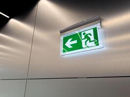 Glowing green emergency exit sign with arrow and running person symbol on brushed metal wall. Evacuation indicator in modern public building. Concept of alert, escape route and safety compliance photo