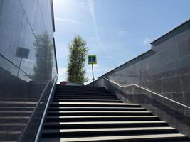 Stairway leading from underground passage to street level with pedestrian sign at top, surrounded by dark stone walls and stainless steel handrail, captured in bright daylight with strong reflections photo