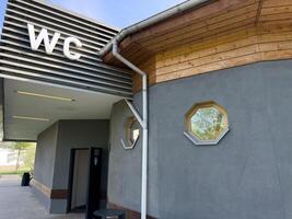 Contemporary public restroom with large WC sign, open door entrance, wooden roof detail and octagonal windows, combining practical accessibility with clean architectural elements in park environment photo