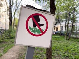 Warning sign in urban park prohibiting walking on the grass. Signpost with red circle and black boot pictogram placed in a green public area surrounded by trees and buildings photo