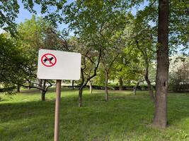 Sign with no dogs allowed symbol on white background mounted on post in green park area with trees and grass, representing public restriction and pet free zone in natural urban environment photo