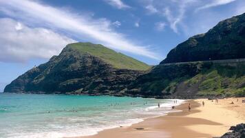 A tropical beach with turquoise waters surrounded by towering green cliffs and sunbathers relaxing on golden sands under video