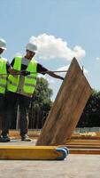 Construction Workers Positioning Large Board on Site. Two construction workers in high-visibility vests and hard hats work together to position a large wooden video
