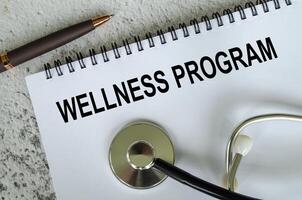 Top view of Wellness Program text on white paper notepad with stethoscope on wooden table background. Health and medical concept. photo