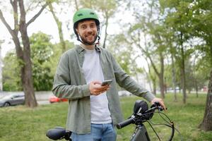 Smiling man in green helmet enjoys cycling in park while using smartphone on sunny day photo