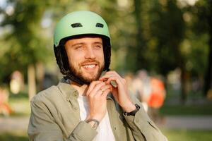 Young man adjusts green helmet while smiling in a sunny park setting during daytime photo