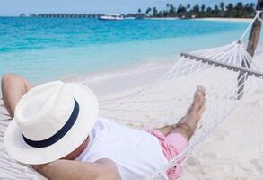 A young man in shorts and a T-shirt is lying in a hammock on the beach. A tourist in a hat enjoys a vacation on tropical islands. photo