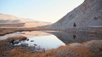 Static back view slow motion adventurous fit traveller cycle bicycle touring solo in remote high Pamir mountains early morning by river. Pamir highway Bartang valley video
