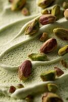 Macro photo of pistachio gelato with whole nuts scattered on the surface. The creamy light green texture features soft wavy strokes and ridges made by a scoop.