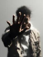 A person with curly hair and neutral-colored clothes is standing behind a textured, frosted glass panel, pressing their hand against it. photo