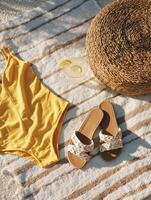 Top-down view on a light striped blanket lies a vibrant yellow one-piece swimsuit, next to a pair of summer sandals with braided details and white straps. photo