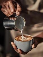 Close-up of a barista pouring milk into a latte, creating a leaf-shaped latte art. The hands frame the image, focus is on the milk stream and coffee surface. photo