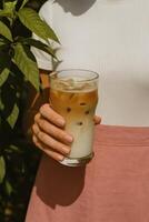 A close-up of a woman holding a glass of iced latte with milk and espresso swirls, visible ice cubes, and a gentle gradient from creamy white to coffee brown. photo